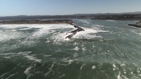 Pulling-back-as-ocean-waves-crash-on-the-narrow-Coquille-River-jetty,-Oregon,-aerial
