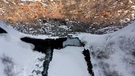 Lowering-aerial-beside-steep-rock-wall-with-river-surrounded-by-white-snow