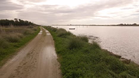 4K-aerial-view-of-a-dirty-road-in-the-bedside-of-Ria-de-Aveiro-in-the-estuary-of-river-Vouga,-drone-moving-forward-toward-a-boat-silhouette,-60fps