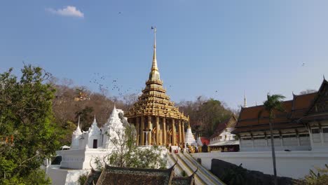 Wat-Phra-Phutthabat,-Saraburi,-Thailand,-steady-aerial-shot-of-the-golden-Buddhist-temple,-people-worshipping,-pigeons-flying-around-in-flocks,-brown-dying-trees-at-the-background-then-a-blue-sky