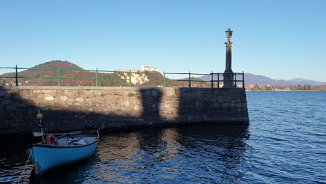 Boot,-Das-Auf-Dem-Wasser-Des-Lago-Maggiore-Am-Pier-Von-Arona-Mit-Der-Festung-Angera-Im-Hintergrund-Schwankt