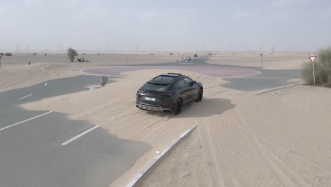 Black-Lamborghini-Urus-drives-across-sand-dune-on-desert-road-in-Dubai,-UAE