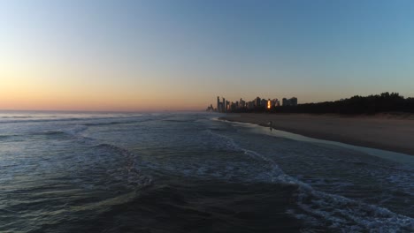 Luftaufnahme-Eines-Wunderschönen-Sonnenaufgangs,-Der-Die-Stadt-Des-Oberflächenparadieses-Im-Hintergrund-Beleuchtet,-Mit-Einem-Surfer,-Der-Ins-Wasser-Eindringt,-Um-Einige-Wellen-An-Der-Southport-Gold-Coast-Qld-Australia-Zu-Fangen
