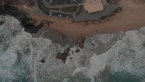 Aerial-top-down-static-shot-over-rough-ocean-and-waves-crashing-on-beach,-Ponta-da-Calheta