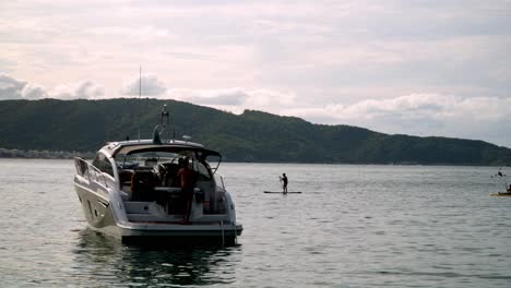 People-relaxing-in-a-yacht-while-others-are-kayaking-in-Praia-da-Sepultura-sea-with-mountains-in-background,-Brazil