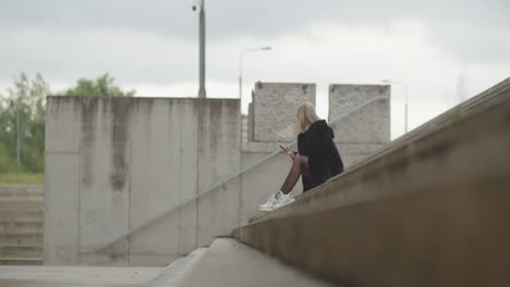 Junge-Blonde-Frau,-Die-Draußen-An-Der-Treppe-Sitzt-Und-Tagsüber-Das-Smartphone-überprüft