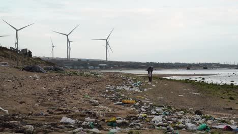 Persona-Caminando-Por-Una-Playa-Contaminada-Con-Un-Parque-Eólico-Visto-Al-Fondo-En-Son-Hai,-Vietnam