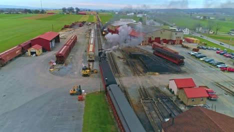 Una-Vista-Aérea-De-Un-Antiguo-Tren-De-Vapor-Restaurado-Y-Vagones-De-Pasajeros-Que-Se-Acercan-Con-Humo-Y-Vapor-Tirando-Hacia-El-Patio-Y-La-Estación-Mientras-Un-Segundo-Tren-De-Vapor-Se-Va-Visto-Por-Un-Dron