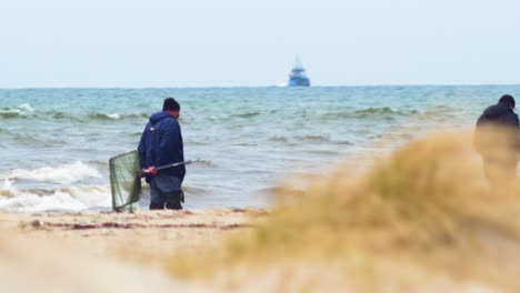 Zwei-Männer-Mit-Fischnetz,-Die-An-Bewölkten-Frühlingstagen-Natürliche-Bernsteinsteine-An-Der-Ostseeküste-Suchen,-Fischerboot-Im-Hintergrund,-Hitzewellen,-Mittlerer-Schuss-Aus-Der-Ferne