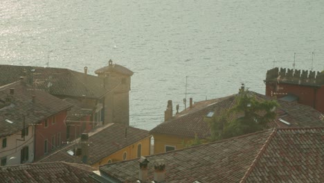 Ganzaufnahme,-Malerischer-Blick-Auf-Das-Dach-Und-Die-Schornsteine-Von-Malcesine-In-Italien,-Die-Sonne-Geht-Auf-Und-Reflektiert-Den-Lago-Di-Garda-Im-Hintergrund