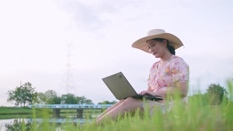 Schöne-Asiatische-Frau-Mit-Hut,-Die-Auf-Dem-Rasen-Sitzt-Und-Am-Computer-Arbeitet-Und-Sich-Erfolgreich-Fühlt