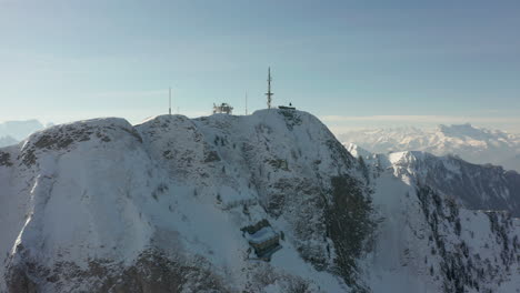 Drone-flying-towards-radio-tower-on-top-of-snow-covered-mountain-summit-and-revealing-beautiful-valley