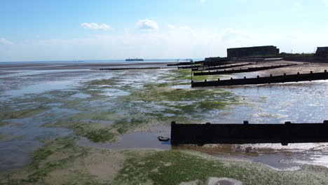 Un-Dron-En-Ascenso-De-Una-Guarnición-Militar-Abandonada-Situada-Detrás-De-Un-Malecón-En-El-Borde-De-La-Playa-Y-Marismas-En-Un-Estuario-De-Marea-Con-Marea-Baja,-En-Un-Día-De-Verano-Claro-Y-Caluroso