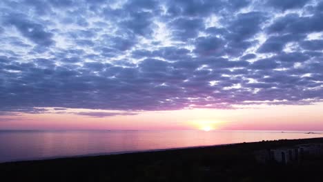 Beautiful-aerial-vibrant-high-contrast-pink-purple-sunset-with-blue-clouds-over-Baltic-sea-at-Liepaja,-distant-ships-in-the-sea,-wide-angle-ascending-drone-shot-camera-tilt-down-slow-zoom-in