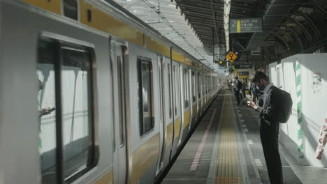 Joven-Con-Mascarilla-Con-Mochila-Usando-Un-Teléfono-Inteligente-En-La-Plataforma-De-La-Estación-De-Tren-En-Tokio,-Japón