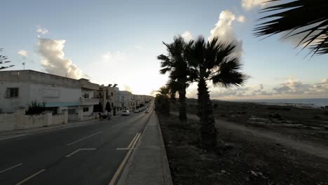 Malta,-Pembroke,-Atemberaubender-Sonnenuntergang-über-Einer-Außenstraße-Und-Bürgersteig-Mit-Palmen-Und-Pendelnden-Autos-Und-Menschen