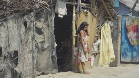 Pobre-Niña-Asiática-De-Piel-Oscura-Sale-De-Su-Casa-De-Campo-Descalza-Y-Mira-Afuera,-Zona-De-Tugurios