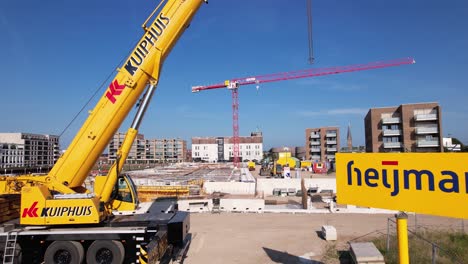 Acercamiento-Aéreo-Lento-Y-Paso-Entre-El-Cartel-Con-El-Nombre-De-La-Empresa-Y-Maquinaria-Pesada-Que-Revela-El-Sitio-De-Construcción-Del-Complejo-Kade-Zuid-De-Apartamentos-De-Lujo-Que-Se-Está-Construyendo-Con-Una-Gran-Grúa-Roja-En-El-Fondo