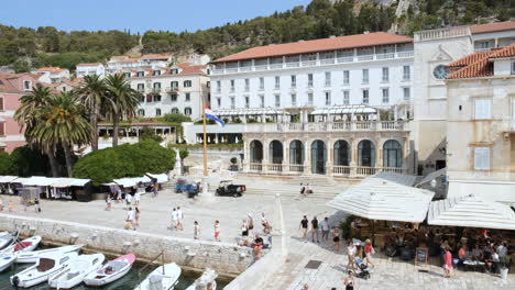 Vista-Aérea-De-Turistas-Caminando-Por-El-Puerto-De-Hvar-Con-Restaurantes-Y-Edificios-Frente-Al-Mar-En-Croacia