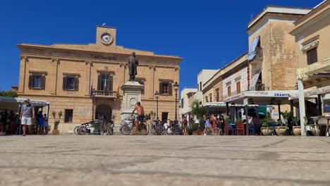 Tiefwinkel-Des-Mannes-Im-Fahrrad-Auf-Der-Piazza-Europa-Auf-Der-Insel-Favignana-Mit-Rathaus-Im-Hintergrund,-Sizilien