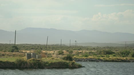Peaceful-telephoto-footage-of-a-pond-and-telephone-poles-going-into-the-distance,-large-clouds-and-mountains-are-seen-in-the-distance,-with-sagebrush-and-other-dry-grasses