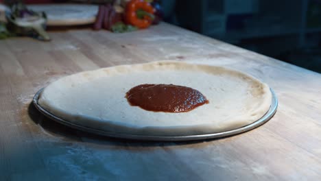pizza-dough-on-a-steel-tray-on-wooden-counter-while-an-expereinced-chef-put-red-sauce-on-it-with-a-adicid