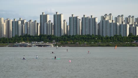 Several-People-Enjoying-Paddle-Boarding-Sports-At-Han-River-With-Jamshil-Apartment-Complex-In-Seoul,-South-Korea