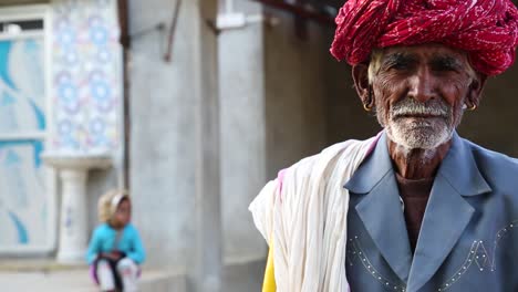 Nahaufnahme-Eines-Alten-Indischen-Seniors-Mit-Rotem-Turban,-Der-Sich-Auf-Einen-Gelben-Stock-Stützt,-Und-Ein-Kind,-Das-Im-Hintergrund-Auf-Der-Treppe-Sitzt