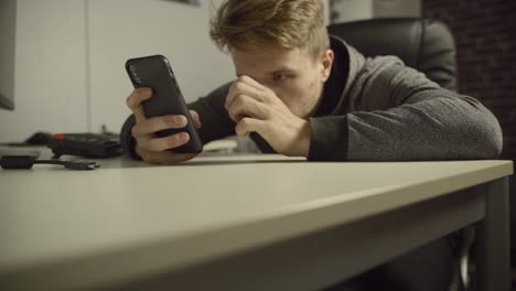 A-young-adult-man-sits-at-his-desk-as-he-uses-his-mobile-phone