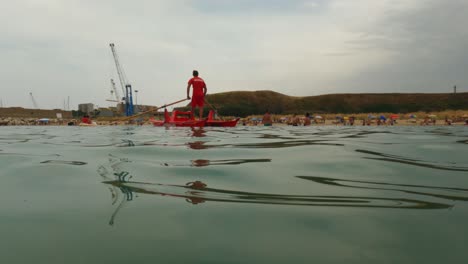 Roter-Italienischer-Rettungsschwimmer-Bereit-Für-Erste-Hilfe-Notfälle-Im-Stehen-Und-Rudern-Von-Rettungsbooten,-Während-Er-Die-Menschen-Beim-Schwimmen-Und-Strand-Im-Hintergrund-In-Punta-Penna-In-Italien-Beobachtet