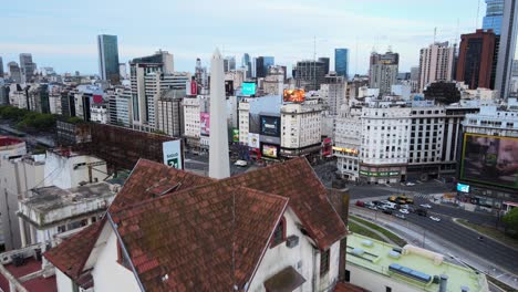 Busy-downtown-Buenos-Aires-with-flashy-billboard-advertisings,-drone-reverse-flying-away-from-monumental-obelisk,-reveals-cute-little-chalet-house-muebleria-diaz-perched-on-top-of-a-terrace-building