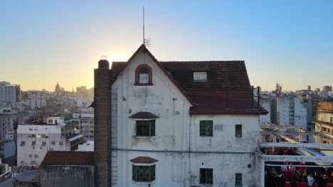 Spectacular-shot,-drone-forward-flying-over-norman-style-little-chalet-house-of-muebleria-diaz-toward-the-big-bright-sun,-slowly-setting-below-the-horizon-with-dense-urban-cityscape-view