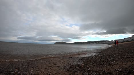 Time-lapse-waves-pulse-across-gloomy-pebble-beach-with-couple-walking-down-Llandudno-coastline