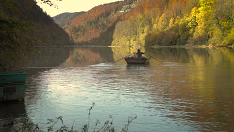 Slow-motion,-man-rowing-boat-on-lake-during-autumn,-surrounded-by-red-yellow-autumn-foliage