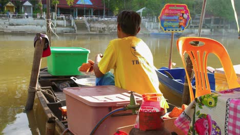 Hombre-Tailandés-De-Espaldas-A-La-Cámara-Friendo-Comida-Callejera-Típica-Asiática-A-Orillas-Del-Río-En-Un-Mercado-Flotante