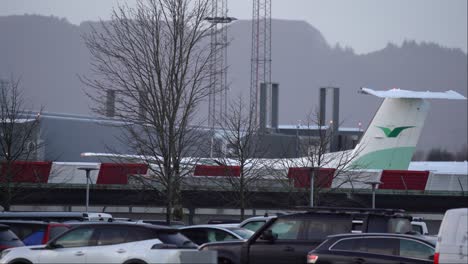 Propeller-airplane-from-Wideroe-company-starting-up-and-reversing-out-from-parking-spot-at-Flesland-airport-Bergen-Norway---Static-with-parked-cars-in-foreground