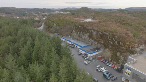 Rema1000-grocery-store-in-Skogsvaag-Oygarden-Norway---Aerial-approaching-building-with-green-forest-in-foreground