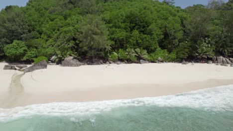 Mahe-Seychelles,-reveal-drone-shot-of-Intendance-beach,-palm-trees,-rock-boulders,-people-on-the-beach