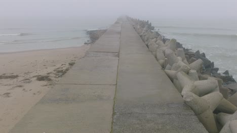 Aerial-establishing-view-of-Port-of-Liepaja-concrete-pier,-Baltic-sea-coastline-,-foggy-day-with-dense-mist,-moody-feeling,-big-storm-waves-splashing,-wide-drone-shot-moving-forward,-tilt-down