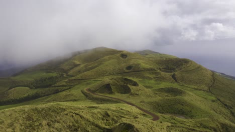 Drone-Dolly-Vista-De-Montañas-Volcánicas-Cubiertas-De-Exuberante-Vegetación-Verde-Con-Camino-De-Tierra,-Nubes-Bajas-Moviéndose-En-La-Isla-De-São-Jorge,-Las-Azores,-Portugal