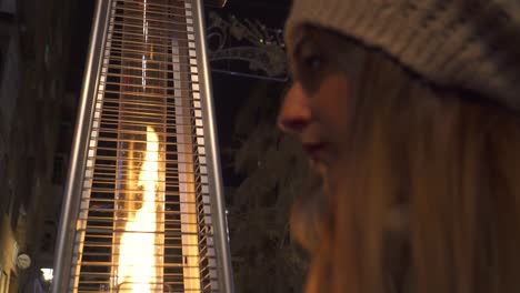 Woman-with-a-hat-placing-her-hair-next-to-an-outdoor-stove-that-gives-off-a-flame-of-fire-and-light