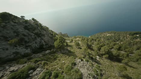 Drone-Aéreo-Fpv-Tiro-Hacia-Adelante-En-Movimiento-Cuesta-Abajo-La-Montaña-Rocosa-Verde-Que-Conduce-Hasta-El-Mar-Azul-Profundo-En-Mallorca,-España