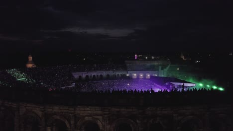 Drone-traveling-on-the-Arenas-de-Nîmes-in-the-middle-of-the-night,-people-are-watching-the-concert-and-there-are-lights-of-several-colors