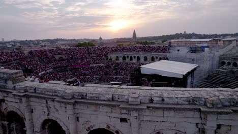 Drone-move-forward-and-up-over-the-Arena-of-Nîmes-at-sunset,-people-are-waiting-for-the-stromae-concert