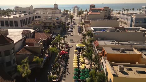 Drohnenvideo-Der-Hauptstraße-In-Huntington-Beach,-Kalifornien,-Die-In-Den-Pazifischen-Ozean-Führt-Und-Den-Berühmten-Pier-Und-Die-Lebhafte-Strandatmosphäre-Mit-Geschäften-Und-Restaurants-Und-Bunten-Sonnenschirmen-Zeigt