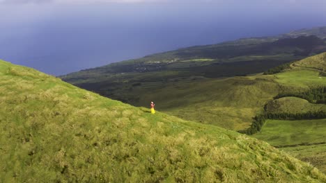 Asian-Malaysian-Chinese-tourist-woman-making-video-or-photo-with-phone-on-a-path-on-the-edge-of-the-vulcanic-green-mountain,-drone-view-on-Pico-da-Esperança,-in-São-Jorge-island,-the-Azores,-Portugal