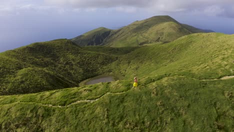 Asian-Malaysian-Chinese-tourist-woman-walking-on-a-path-on-the-edge-of-the-mountain-with-360-camera-selfie-stick,-drone-view-on-Pico-da-Esperança,-in-São-Jorge-island,-the-Azores,-Portugal
