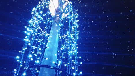 Low-angle-view-of-snow-falling-around-blue-holiday-lights-decorating-light-pole