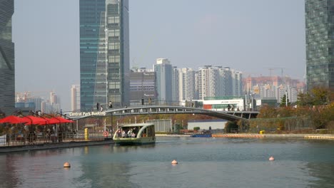 Parque-Central-De-Songdo---La-Gente-Viaja-En-Taxi-Acuático-En-Un-Lago,-Los-Viajeros-Cruzan-Un-Puente-Peatonal-Disfrutando-Del-Horizonte-Urbano-Moderno-Futurista