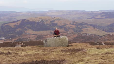 Hombre-Meditando-En-La-Naturaleza-Salvaje-En-Una-Gran-Roca-En-Posición-De-Loto-En-El-Paisaje-Rural-De-Escocia,-Tierras-Altas-Del-Oeste